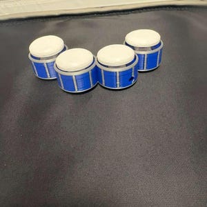 May include: Four small, blue and white containers with a cylindrical shape. Each container has a white top and a blue base with silver accents. The containers are arranged on a dark surface.