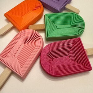 May include: Five colorful popsicle-shaped objects in orange, purple, green, pink, and red. Each has a wooden stick. The popsicle shapes have a layered, ridged design. The objects are arranged on a white surface.