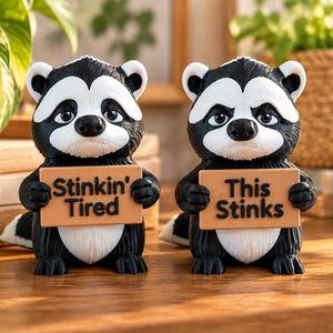 May include: Two black and white skunk figurines standing side by side. The left skunk holds a tan sign that reads Stinkin' Tired. The right skunk holds a tan sign that reads This STINKS. Both figurines have detailed fur textures and white facial markings.
