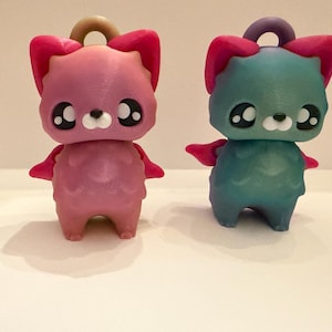May include: Two 3D-printed cat figurines. One is pink with dark pink ears and wings, the other is teal with dark pink ears and wings. Both have large, black-outlined eyes, a white muzzle, and a loop at the top for hanging.