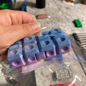 May include: A clear plastic bag holds several blue and purple 3D-printed cube-shaped connectors. Each cube has a hole on each side and a rounded top. The cubes are gradient-colored, transitioning from blue to purple.