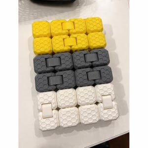 May include: A close-up shot of three connected fidget toys in yellow, gray, and white. Each toy is composed of interlocking hexagonal blocks. The yellow and white toys have a clasp, while the gray ones do not.