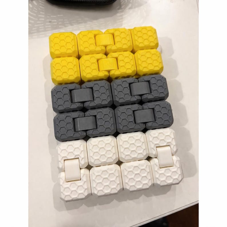 May include: A close-up shot of three connected fidget toys in yellow, gray, and white. Each toy is composed of interlocking hexagonal blocks. The yellow and white toys have a clasp, while the gray ones do not.