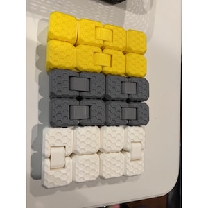 May include: A close-up shot of three rows of interlocking plastic blocks. The top row is yellow, the middle row is gray, and the bottom row is white. Each block has a textured surface and a connecting mechanism.
