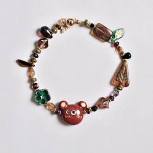 Puede incluir: Un brazalete con un dije de oso marrón con cuentas verdes, marrones y doradas. El brazalete tiene un cierre magnético.