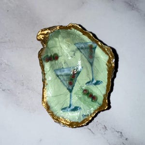 May include: A decorative oyster shell painted with a light green background and gold trim. The shell features two martini glasses with olives. A unique, handcrafted piece for home decor.