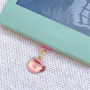 May include: A light green Kindle e-reader with a small pink plastic clip attached to the bottom edge. A gold-toned metal jump ring connects the clip to a small, enamel-style charm shaped like a light pink coffee mug with a dark pink heart design.