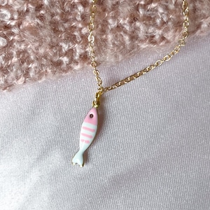 May include: A gold chain necklace featuring a fish pendant. The pendant is pink with white stripes and a light blue tail. The background is a soft pink, textured fabric. The necklace is a delicate piece of jewellery.