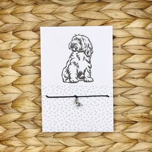 May include: A black string bracelet with a silver charm of a dog. The bracelet is packaged on a white card with a black and white illustration of a dog.