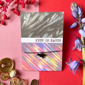 May include: A black raven charm on a purple string bracelet with the words "KEEP ON RAVEN" on a card with a feather pattern.
