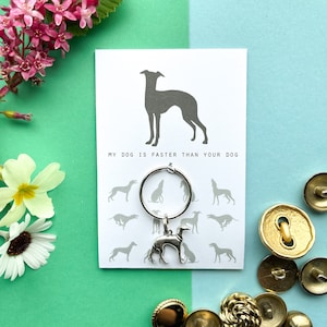 May include: A white card with the text "My dog is faster than your dog" and a silver dog charm attached to a silver ring. The card has a silhouette of a greyhound dog on the top and several smaller greyhound silhouettes around the text. The card is surrounded by pink and yellow flowers and gold buttons.