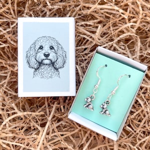 May include: A pair of silver-toned metal dog-shaped dangle earrings with hook backings, presented inside a small white box with a light green insert. The box lid features a detailed black and white line drawing of a curly-haired dog face.