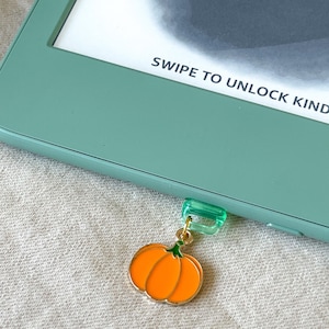 May include: A pumpkin-shaped charm in orange with a gold border and green stem, suspended from a green clip. The clip is attached to a mint-green e-reader. Text reads "SWIPE TO UNLOCK KINDLE".