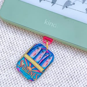 May include: A light green e-reader with a screen displaying birds on wires, attached to a rectangular acrylic charm featuring a colourful illustration of sardines in a tin with the text SARDINES PURE OLIVE OIL, connected by a small pink plastic loop.