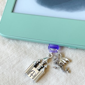 May include: Silver charms of a castle and a dragon suspended from a purple clip. The clip is attached to a mint-green e-reader.