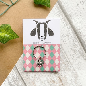 May include: A silver charm featuring a goat with a pink and green argyle pattern background. The charm is attached to a silver ring. The card reads "Seasons Bleatings".