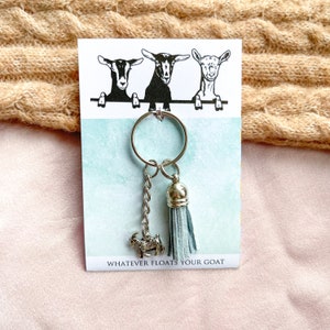 May include: A silver keychain with a blue tassel and a goat charm. The keychain is attached to a card with three goats on it and the text "Whatever floats your goat".