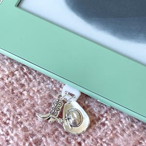 May include: Silver-coloured cowboy boot and hat charms suspended from a white clip. The charms feature detailed designs. The background includes a light green frame and a pink, textured surface.