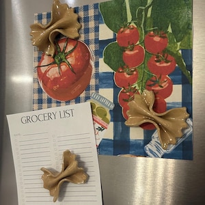 May include: A fridge magnet set with a grocery list and decorative magnets. The magnets are shaped like pasta bows and are tan. The grocery list is white with the words "GROCERY LIST" at the top. The background includes images of tomatoes.