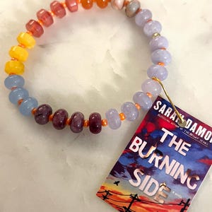 May include: A beaded bracelet featuring a sequence of round, smooth beads in shades of orange, yellow, light blue, lavender, and deep purple, strung on an orange cord. A rectangular tag attached by a gold safety pin displays the text THE BURNING SIDE.
