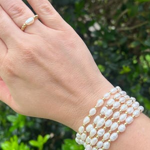 May include: A hand wearing a gold ring with a pearl and a stack of pearl bracelets with gold beads. The jewelry is set against a backdrop of green foliage.
