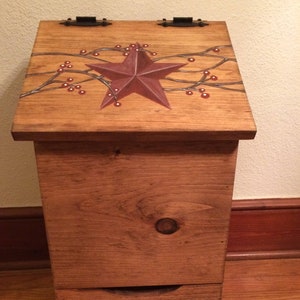 May include: A wooden storage chest with a hinged lid and a drawer. The chest is stained a warm brown and features a large burgundy star and berry branch design on the lid. The drawer has three burgundy stars and berry branches.