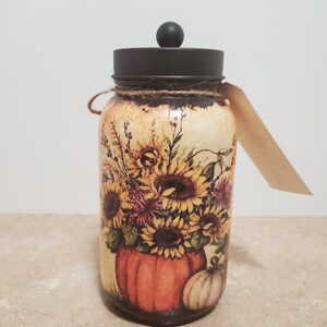 K&ouml;nnte beinhalten: Ein Glasgef&auml;&szlig; mit einem schwarzen Deckel und einem Herbstmotiv. Das Gef&auml;&szlig; zeigt einen K&uuml;rbis, Sonnenblumen und andere Herbstlaub in Orange-, Gelb- und Braunt&ouml;nen.