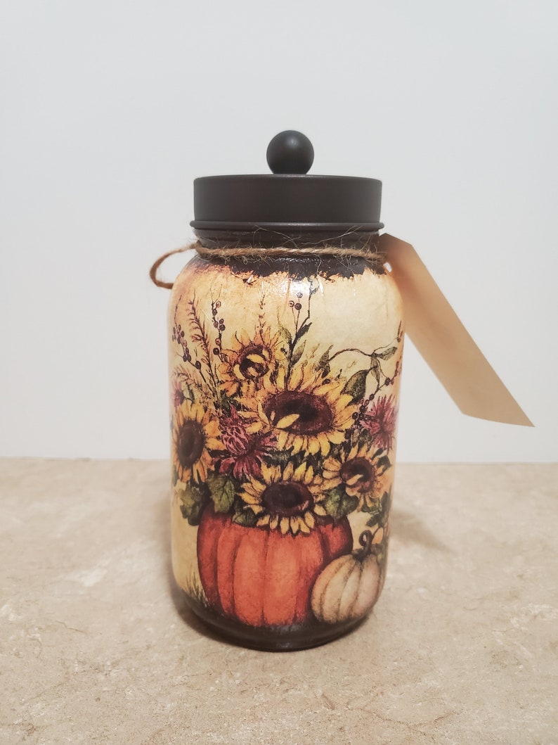 K&ouml;nnte beinhalten: Ein Glasgef&auml;&szlig; mit einem schwarzen Deckel und einem Herbstmotiv. Das Gef&auml;&szlig; zeigt einen K&uuml;rbis, Sonnenblumen und andere Herbstlaub in Orange-, Gelb- und Braunt&ouml;nen.