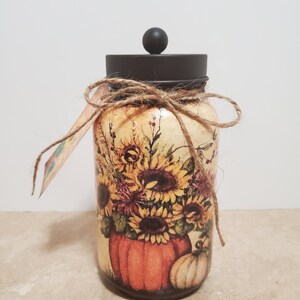 K&ouml;nnte beinhalten: Ein Glasgef&auml;&szlig; mit einem schwarzen Deckel und einer braunen Kordelschleife. Das Gef&auml;&szlig; ist mit einem Herbstmotiv verziert, das Sonnenblumen, K&uuml;rbisse und Bl&auml;tter zeigt.