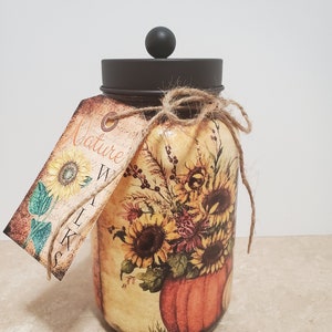 K&ouml;nnte beinhalten: Ein Glasgef&auml;&szlig; mit braunem Deckel und einem Etikett mit der Aufschrift "Nature Walk" ist mit einem Herbstmotiv verziert, das Sonnenblumen, Bl&auml;tter und einen K&uuml;rbis zeigt.