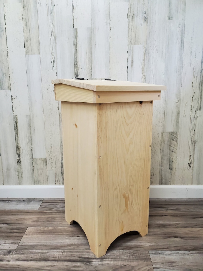 May include: A natural wood bin with a hinged lid and a slightly curved base. The bin is made of unfinished wood and has a simple, rustic design.