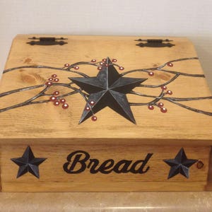 May include: Wooden bread box with a black star and red berries painted on the lid. The front of the box has the word "Bread" written in black cursive and two black stars on either side.