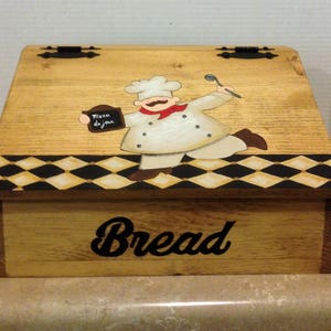 May include: Wooden bread box with a hinged lid. The lid features a painted design of a chef holding a menu that says "Menu du jour". The chef is wearing a white chef's hat, a white chef's coat, and a red scarf. The box is painted with a black and white checkered pattern. The word "Bread" is painted on the front of the box.
