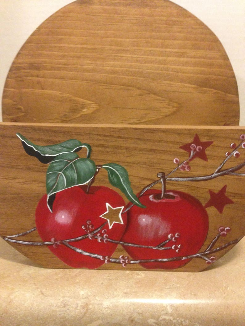 May include: A wooden wall-mounted mail holder with a half-circle top and a painted design of two red apples with green leaves and white stars on a branch with small pink flowers.