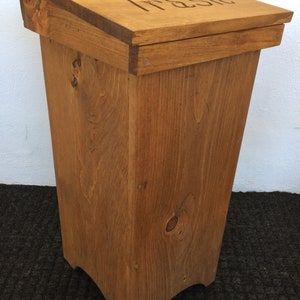 May include: A wooden trash can with a hinged lid and the word "Trash" carved into the top. The can is stained a dark brown color and has a slightly curved base.