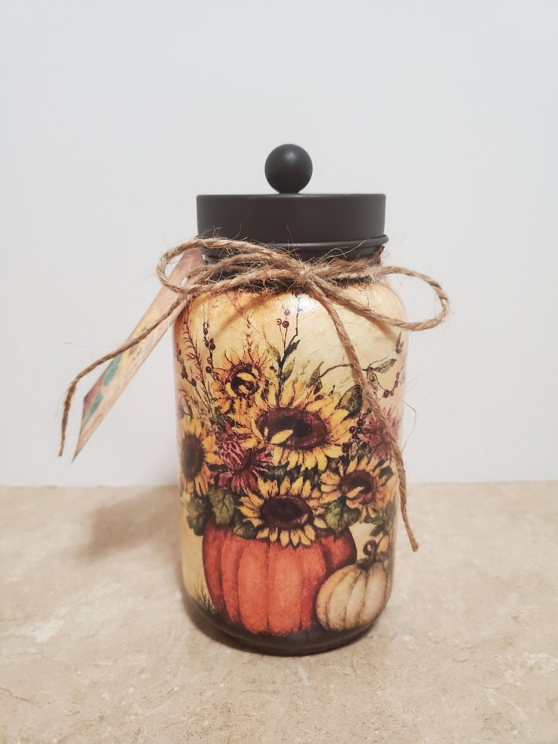 K&ouml;nnte beinhalten: Ein Glasgef&auml;&szlig; mit schwarzem Deckel und einer rustikalen Kordelschleife. Das Gef&auml;&szlig; ist mit einem Herbstmotiv verziert, das Sonnenblumen, K&uuml;rbisse und Bl&auml;tter zeigt.