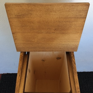 May include: A wooden storage bin with a hinged lid. The bin is made of natural wood with a light brown stain. The lid is open, revealing the empty interior of the bin.