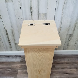 May include: A natural wood storage bench with two black metal hinges on the top. The bench has a simple design with a rectangular top and a slightly curved base.