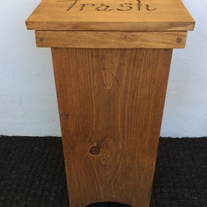 May include: A wooden trash can with a hinged lid. The lid has a cutout for a trash bag and the word "Trash" is carved into the top. The can is stained a dark brown color.