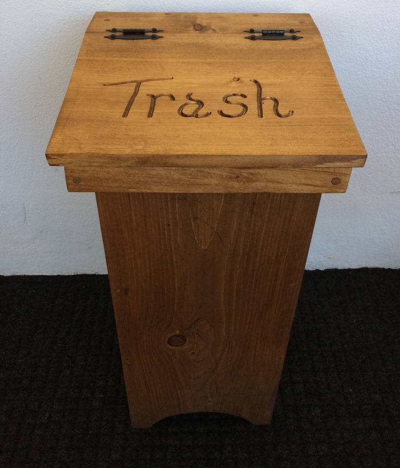 May include: A wooden trash can with a hinged lid and black metal hinges. The lid is engraved with the word "Trash".
