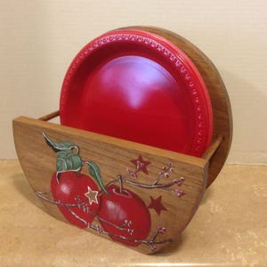 May include: Wooden plate holder with a red apple design. The holder is painted with red apples, green leaves, and brown branches. The holder has a slot for holding plates.