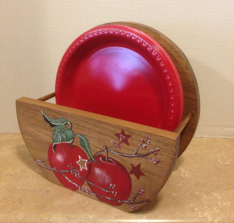 May include: Wooden plate holder with a red apple design. The holder is painted with red apples, green leaves, and brown branches. The holder has a slot for holding plates.