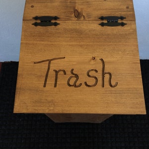 May include: A wooden trash can with a hinged lid and black metal hinges. The lid is closed and the word "Trash" is carved into the wood.