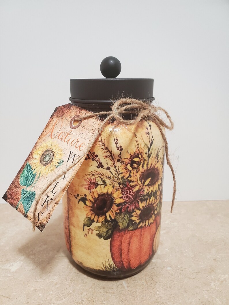 K&ouml;nnte beinhalten: Ein Glasgef&auml;&szlig; mit braunem Deckel und einem Etikett mit der Aufschrift "Nature Walk" ist mit einem Herbstmotiv verziert, das Sonnenblumen, Bl&auml;tter und einen K&uuml;rbis zeigt.