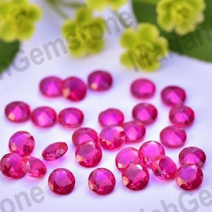 May include: A collection of faceted, round, and oval-shaped pink gemstones. The gems are scattered on a white surface, with a few green leaves and yellow flowers in the background. The gems are likely rubies.