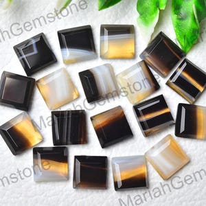 May include: A collection of square-cut gemstones featuring banded patterns in shades of black, translucent white, and amber. Each stone has a smooth, polished surface with distinct horizontal or diagonal layers of colour visible throughout the set.