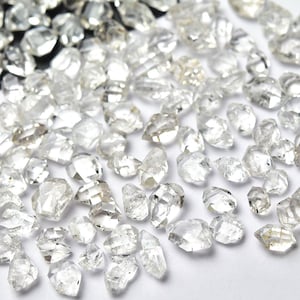 Peut inclure: Une collection de pierres précieuses claires et facettées dispersées sur une surface blanche. Les pierres précieuses varient en taille et en forme, avec de multiples facettes captant la lumière. L'ensemble est un affichage étincelant de diamants bruts.