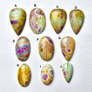 May include: Ten polished cabochon gemstones labeled A through J, featuring a mix of olive green, tan, and vibrant magenta-pink inclusions. The stones are shaped in various ovals and teardrops, displaying natural swirling patterns and smooth surfaces.