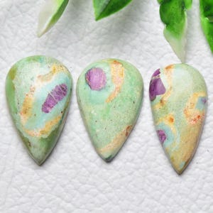 Peut inclure: Trois cabochons en forme de goutte d'eau présentant une surface lisse et polie avec un motif marbré composé de vert menthe clair, de beige et d'inclusions magenta vif. Chaque pierre affiche une répartition unique de ces couleurs.