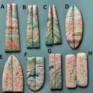 Puede incluir: Surtido de ocho pares de cabujones de piedras preciosas pulidas de varias formas. Las piedras muestran una mezcla de colores, incluyendo rojo, verde y crema, con patrones únicos. Los cabujones se exhiben sobre un fondo verde azulado.